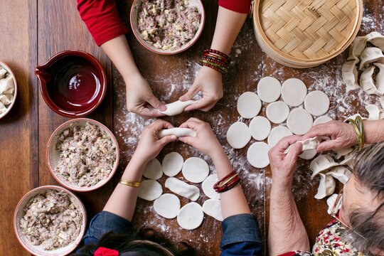 Overhead view of three generations of hands folding dumplings together on a floury wooden table with filling bowls