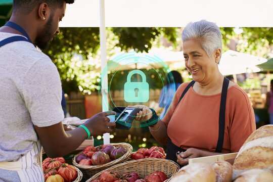 Smiling senior woman vendor accepting secure contactless payment from customer at outdoor farmers market with padlock security icon