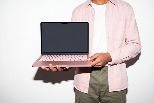 A man in an open pink shirt, white tee and olive cargo pants balances a laptop on his palm, steadying it with his other hand. The blank display points toward the viewer. Direct flash shoot.