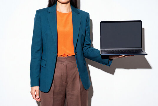 A businesswoman in a teal tailored blazer, orange silk blouse and brown trousers extends an open laptop sideways on her flat palm. The blank display sits in clean white space. Direct flash shoot.