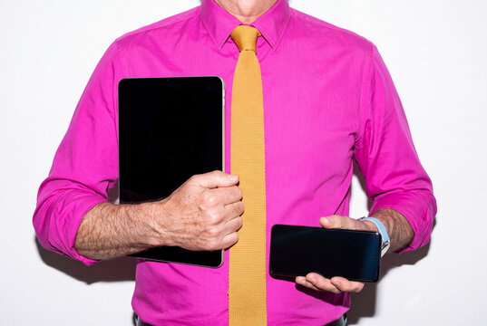 A businessman in a magenta shirt and knitted mustard tie holds a tablet upright in one hand and a smartphone sideways in the other. The bold colors and blank displays suit office mockups. 