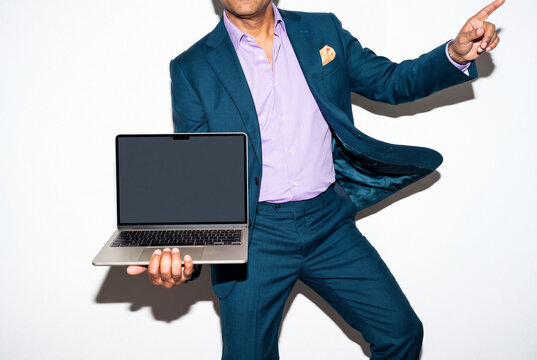 A businessman in a teal suit, lilac shirt and pocket square dances holding an open laptop on one hand and pointing up with the other. His movement conveys celebration and success. Direct flash shoot.