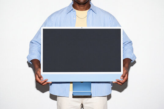 A man in an open striped shirt, pale yellow tee and gold chain holds a blue all-in-one desktop computer in front of his torso. The large blank display is ready for mockups. Direct flash shoot.