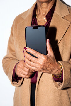 A senior woman in a camel coat and burgundy satin blouse shows a smartphone with both hands, her nails painted to match and layered gold chains at her neck. Direct flash shoot.