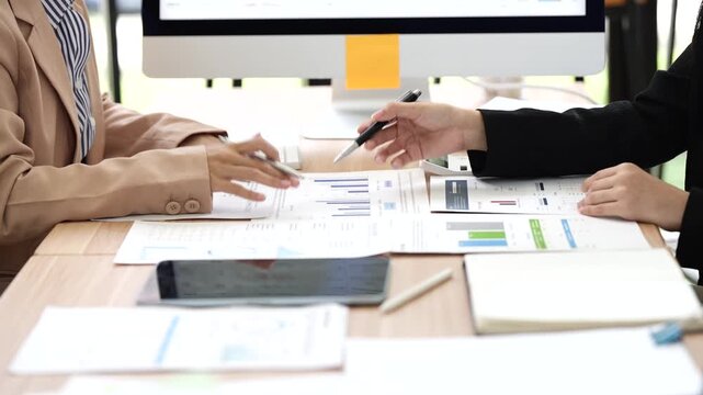 Two businesswomen discussing financial charts and documents at office desk with computer monitor displaying data in background.