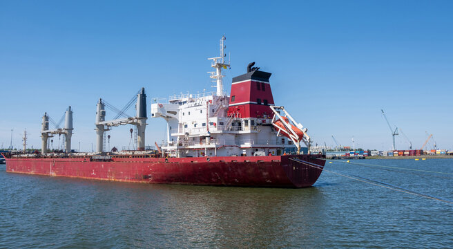 Red bulk carrier with deck cranes moored in Rotterdam harbor, Netherlands, under blue sky