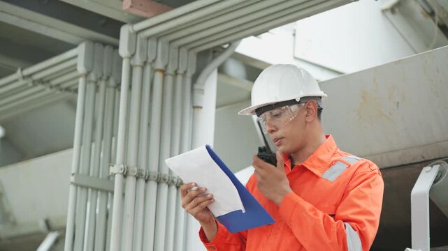 Industrial engineer reviews technical documents on clipboard while inspecting industrial piping system. Facility maintenance, quality control, safety assessment.