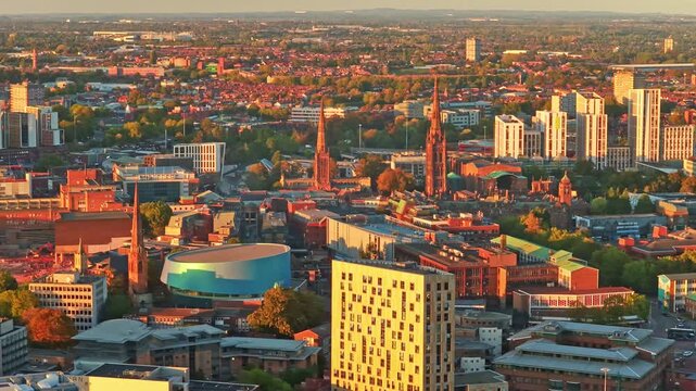 Coventry spires, the round blue building and tower blocks of the city centre glowing at golden hour in the West Midlands, England, aerial