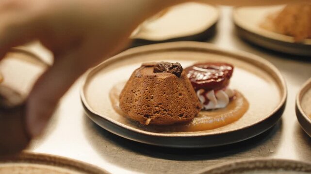 Cook's fingers setting a small chocolate bundt cake onto a swirl of caramel sauce on a stoneware plate, one of a row being plated at a steel kitchen pass.