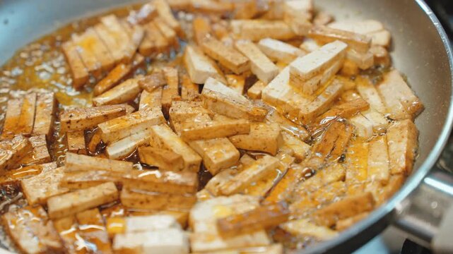 Close view of batons of firm tofu sizzling in oil and soy based sauce in a frying pan, the edges turning golden brown.