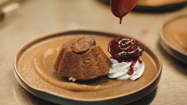 Spoon placing glossy dark plum compote next to a whipped cream rosette and a small chocolate bundt cake on caramel sauce, on a stoneware plate.