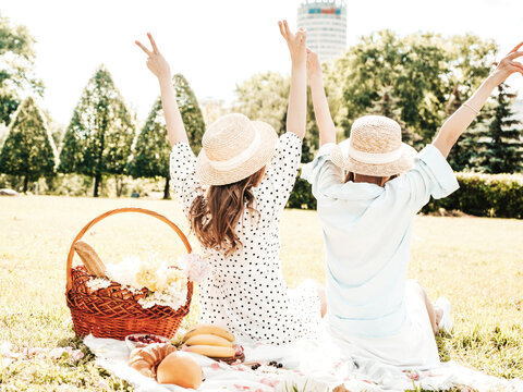 Back view of two young beautiful smiling hipster female in trendy summer sundress and hats.Women making picnic outside.Positive models sitting on plaid on grass, hugging and raising hands