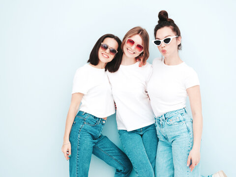 Three young beautiful smiling hipster female in trendy same summer white t-shirt and jeans clothes. Sexy carefree women posing near light blue wall in studio. Cheerful and positive models having fun