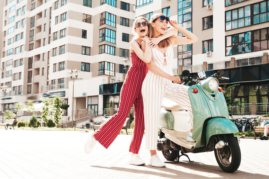 Two young beautiful smiling hipster female in trendy overalls.Sexy carefree women driving retro motorbike on the street background.Positive models having fun, riding classic Italian scooter in eyewear