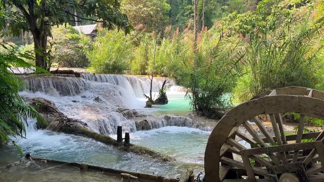 Wooden waterwheel beside tiered cascades and turquoise pools at Kuang Si Waterfall near Luang Prabang, Laos, surrounded by lush tropical vegetation.