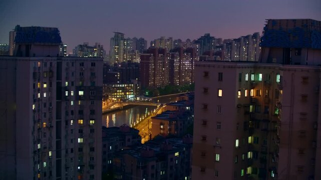 Tall apartment buildings overlook a warmly lit street in Shanghai as evening darkens the sky, with glowing windows and rows of distant housing adding depth to the closely packed residential cityscape.