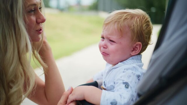Mother soothes crying toddler in stroller. Gentle touch calms upset child. Outdoor setting shows care and patience. Ideal for parenting, childcare, or emotional support themes