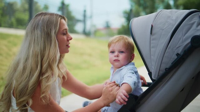 Mother smiles as toddler points finger at her. Child sits in stroller, engaged and curious. Mother gently holds child, showing affection. Outdoor park scene with green grass and trees