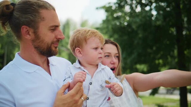 Family of three enjoys outdoor moment together. Man holds toddler securely. Woman smiles beside them. Child points excitedly toward something. Scene shows happy, relaxed family bonding