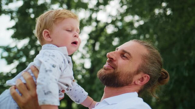 Joyful father lifts his laughing toddler outdoors. The child beams with delight, arms raised. Dad smiles warmly, eyes soft with happiness. Green trees softly blur behind them