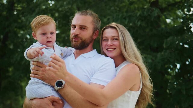 Young family smiles in park. Father holds baby, mother laughs beside. Child points toward something. Natural light, green trees background. Ideal for family, parenting, or outdoor lifestyle themes