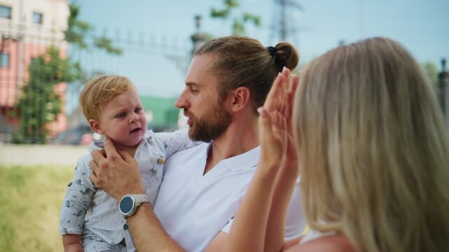 Happy father holds toddler while mother claps joyfully. Parents smile warmly with child. Outdoor park setting with green grass and fence. Ideal for family events, parenting content