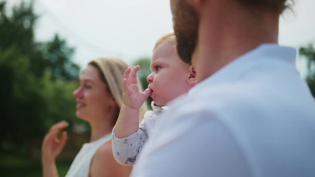Happy family outdoors. Man holds baby while woman smiles. Baby waves hand joyfully. Warm, natural light enhances peaceful moment. Ideal for parenting, family, or lifestyle content
