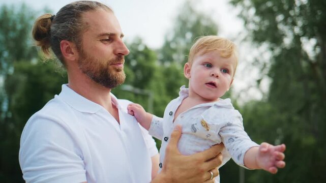Father holds his young child in a park. Father gazes at child with tender affection. Child looks upward, curious and calm. Sunlight filters through green trees around them. Footage suits family
