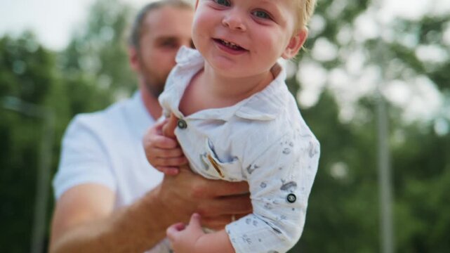 Father holds joyful baby boy in park setting. Man with beard gazes lovingly at child. Bright sunlight enhances tender bonding moment. Ideal for family, parenting, or outdoor lifestyle themes