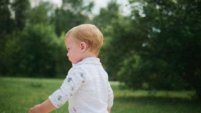 Blond toddler walks outdoors, arms outstretched, focused on movement. Expression shows determination, slight smile visible. Scene captures early childhood exploration in green park