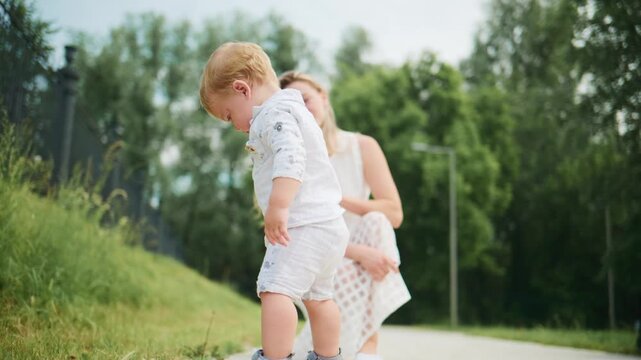 Mother gently lifts her toddler son in a sunny park. Child laughs, enjoying playful moment with mom. Green trees and grass surround them. Scene captures joyful family bonding outdoors