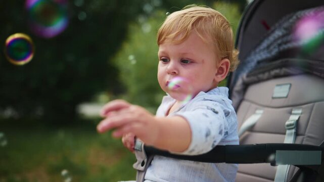 Baby in a stroller watches colorful bubbles float by. The child's focused gaze shows curiosity and wonder. Soft outdoor light highlights gentle exploration. Ideal for advertising baby products