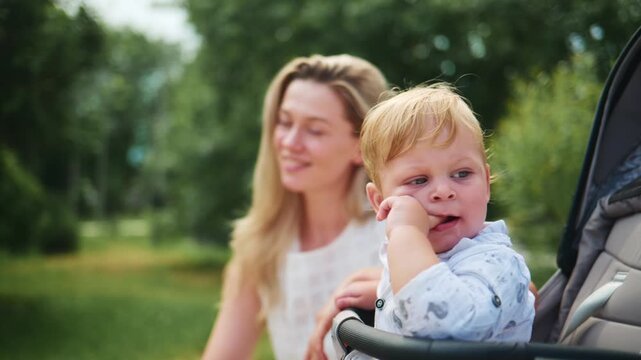 Mother smiles as she pushes stroller through sunny park. Child looks forward with curious expression. Scene captures joyful, relaxed family outing. Perfect for parenting, travel, or lifestyle content
