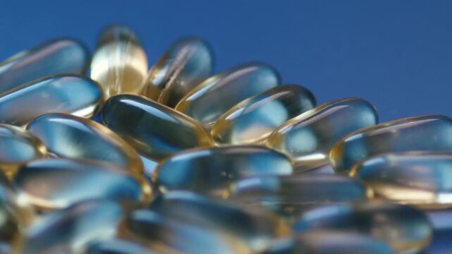 A group of golden gelatine capsules with fish oil rotates on a blue glossy surface. A close-up shot demonstrates the texture and translucent shell of the medical preparations