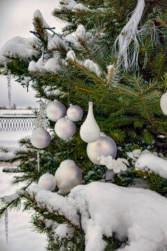 A Christmas and New Year tree stands covered in snow, adorned with various shiny ornaments hanging from its branches near a snowy landscape