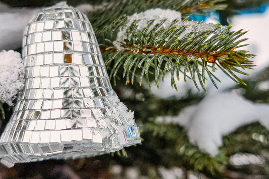 A Christmas and New Year tree is in the snow. A reflective bell ornament hangs on a branch among green needles and snowy patches