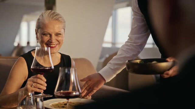 Senior Caucasian woman receiving dish from waiter and toasting with red wine glass during dinner date. Footage fits hospitality advertising, senior dating campaigns, and luxury lifestyle marketing