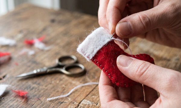 Closeup of hands sewing a small red felt christmas stocking ornament with needle and white thread on a rustic wooden table, surrounded by crafting materials and vintage scissors for diy craft project
