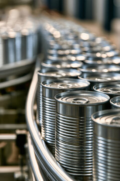 Ribbed tin cans with ring-pull lids are in motion on a conveyor belt. The cans closest to the viewer are sharp in focus while others fade into the