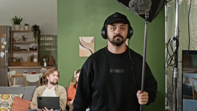 Portrait of Caucasian young adult sound technician holding boom microphone on indoor set. Useful for film industry promotions, audiovisual courses, and media crew training