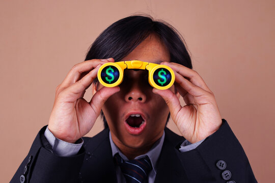 Surprised Asian Businessman Holding Binoculars with Dollar Symbols on Lenses Over Brown Background