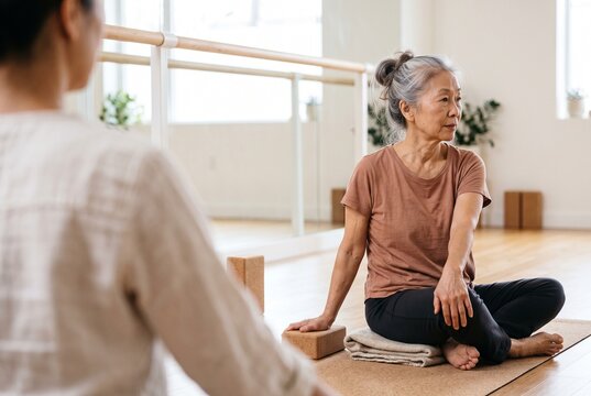 Active senior Asian woman practicing seated spinal twist yoga pose on cork mat with blocks in bright fitness studio during guided wellness class