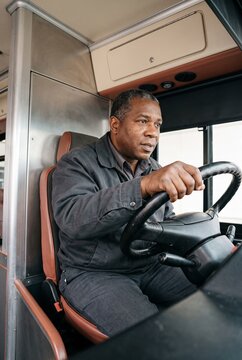 Focused mature black male bus driver sitting in the cabin holding the steering wheel and looking ahead at the road while operating public transport