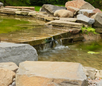Gentle waterfall cascade flowing over flat flagstones into an artificial garden pond surrounded by large natural rocks and boulders.