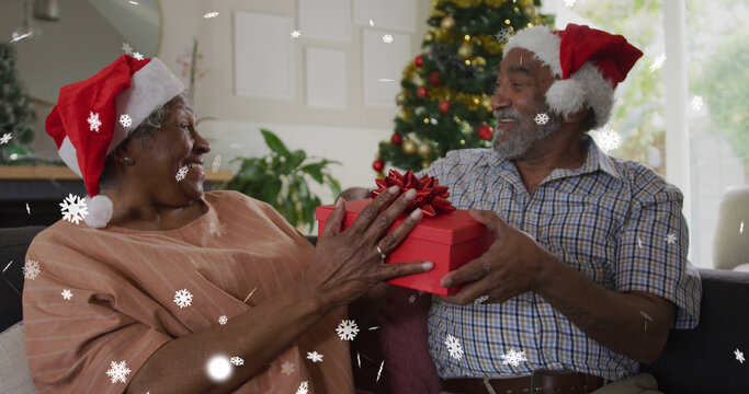 Image of snow falling over senior african american couple wearing santa hats