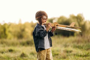 Child wearing vintage aviator goggles and holding a wooden model airplane outdoors in a sunlit grassy field with blurred trees in the background © standret
