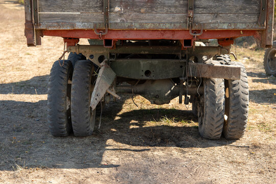 Rear view of an old farm trailer showing its undercarriage, worn dual tires, rusty metal frame and weathered wooden bed. Aging agricultural equipment standing outdoors on dry ground.
