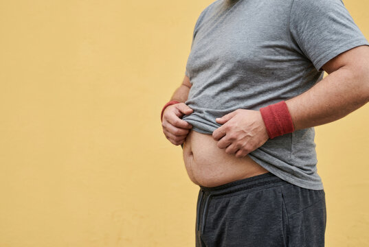 Unhealthy belly fat overweight man male body sport wristband casual clothing yellow wall weight problem unhealthy lifestyle midsection closeup sedentary habit Unhealthy belly fat overweight man