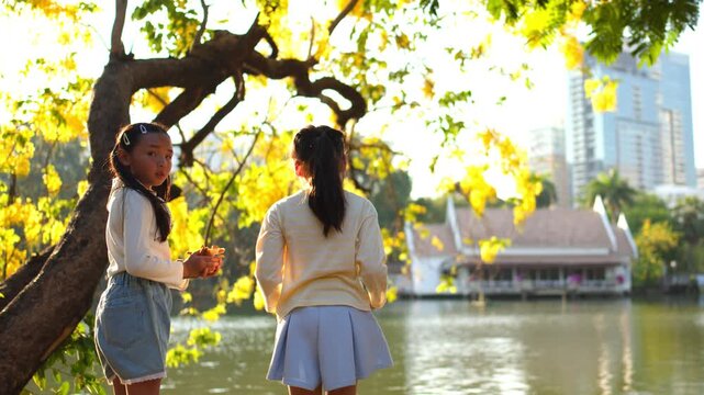 Two Young Asian Girls Standing by a Park Lake Under Flowering Trees