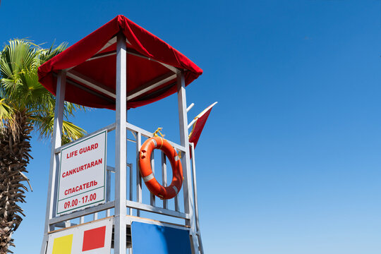 A red-roofed lifeguard observation tower with an orange life buoy ring and multilingual safety sign (English, Turkish, Russian) against a clear blue sky. 